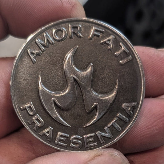 A close-up of a round metallic coin or medallion held in a person's hand. The coin has an aged silver or bronze-toned finish with raised text reading 'AMOR FATI' at the top and 'PRAESENTIA' at the bottom, surrounding a central flame-like emblem.