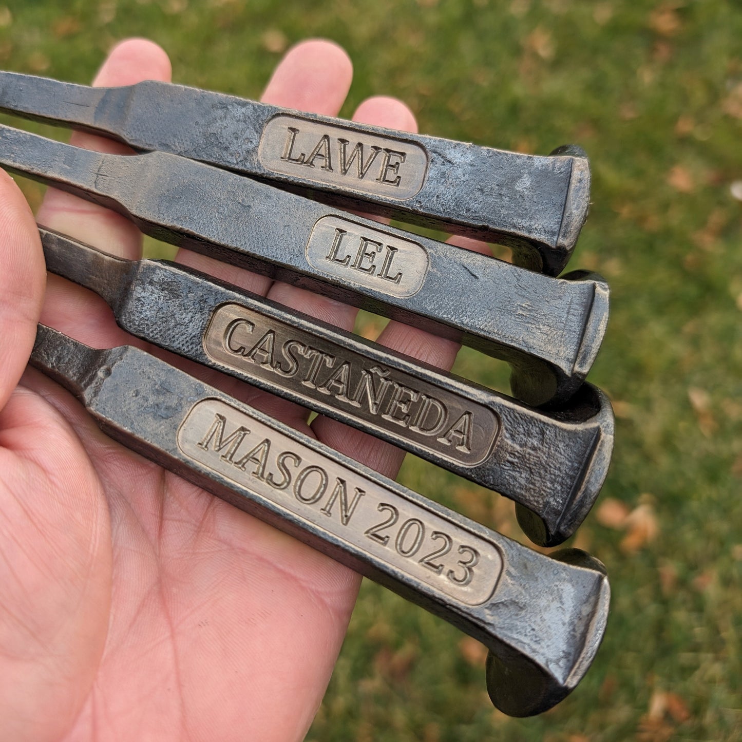 Metal tools with engraved names and years held in a hand against a grassy background