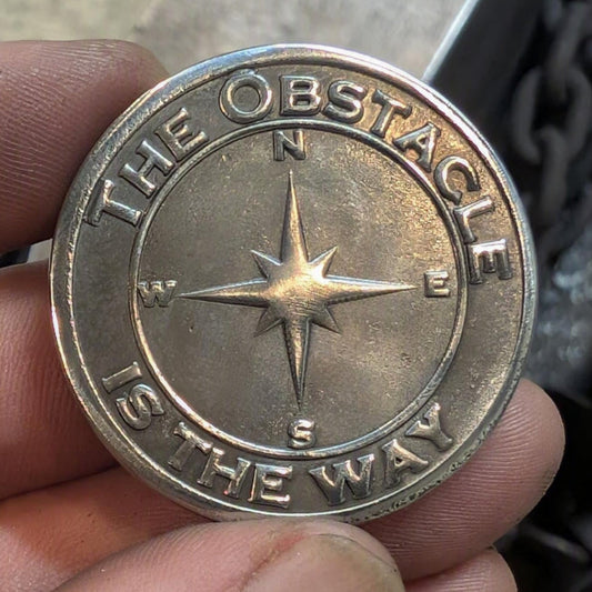 A close-up of a hand holding a round metallic medallion or coin-like token with an embossed compass rose and the text 'THE OBSTACLE IS THE WAY' around the edge.