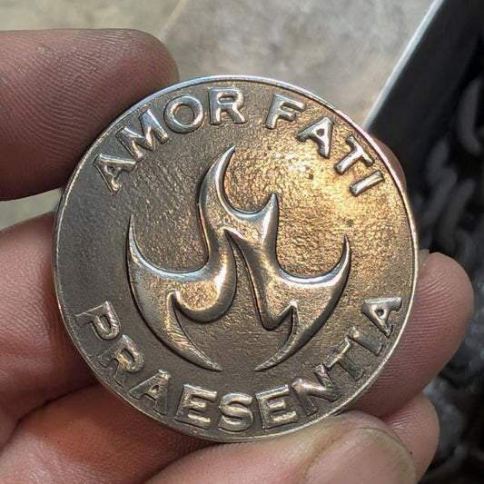 A close-up of a hand holding a round metallic coin or medallion engraved with the words 'AMOR FATI' at the top and 'PRAESENTIA' at the bottom, surrounding a central flame-like emblem. The background shows a blurred workshop or industrial setting.