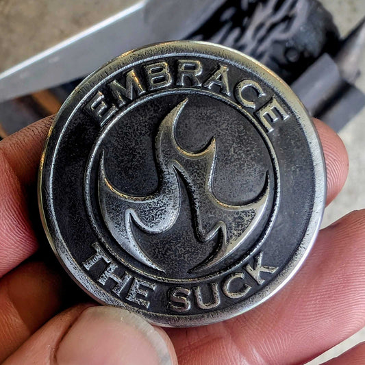 Person holding a metallic coin with 'Embrace the Suck' text and symbol.