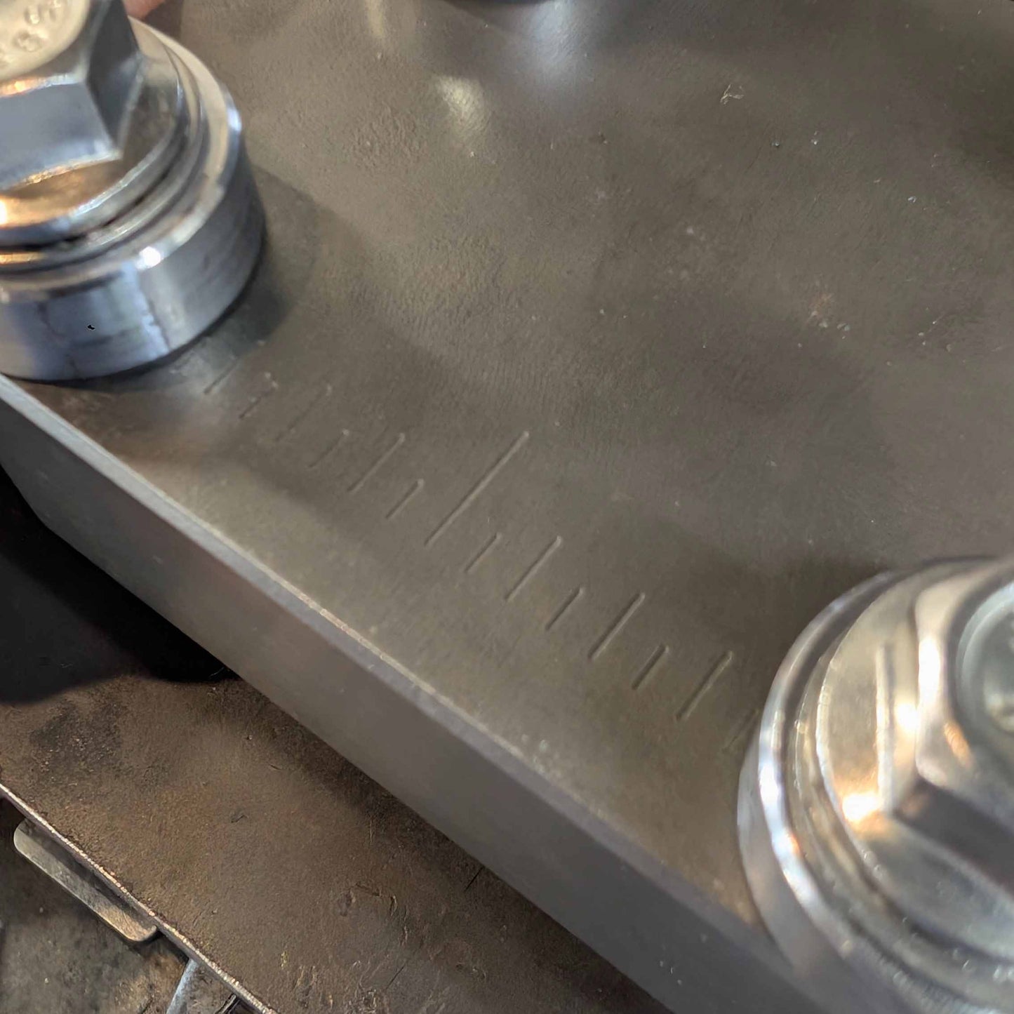 Close-up of a dark gray machined metal surface with machined measurement markings and silver hex nuts attached at the corners.
