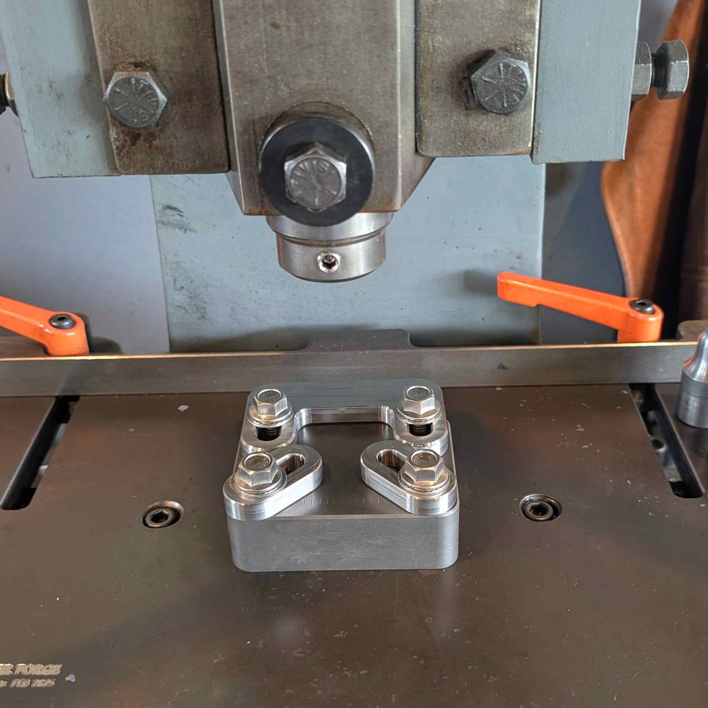 A close-up of a machined metal block with four hex bolts secured in a clamp, positioned on a dark metal work surface under a press or machine with orange adjustment handles.