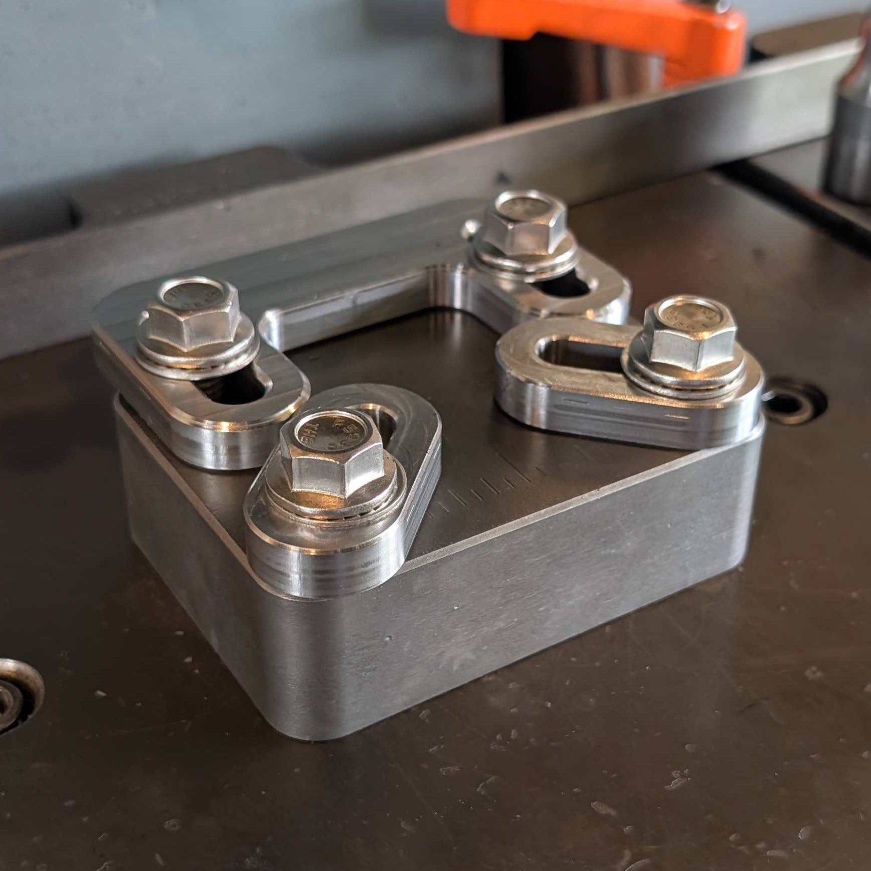 A machined metal mechanical component with four large hex nuts and washers, resting on a dark workbench with a metal bar and orange clamp visible in the background.