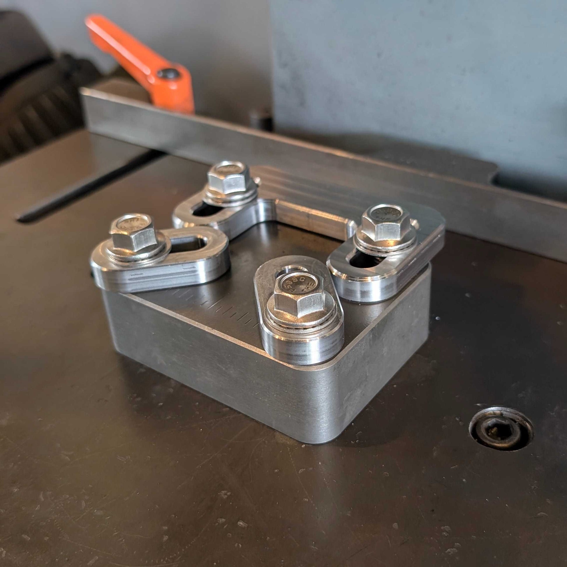 A machined metal mechanical component with four bolted flange connections, resting on a dark workbench with industrial machinery in the background.