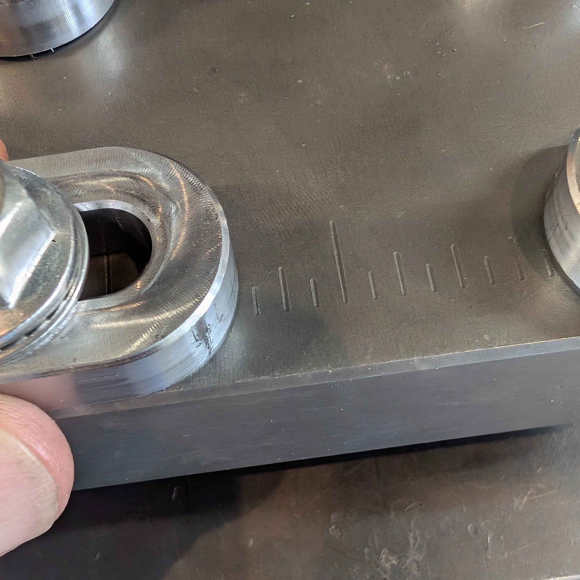 A close-up of a machined metal part with a hexagonal threaded section, held by a hand over a dark metallic work surface with engraved measurement markings.
