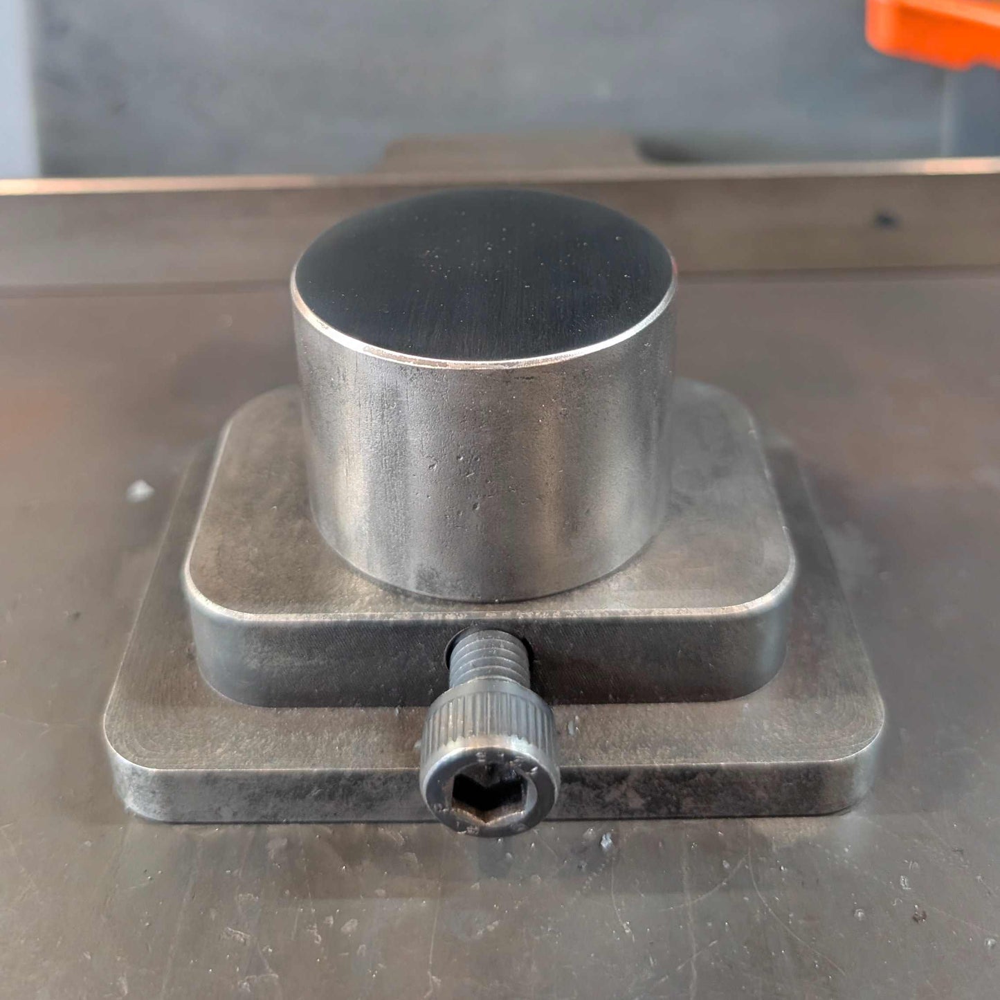 A close-up of a metal cylindrical workpiece resting in a clamp under a large metal press or machine head, with a gray industrial background.