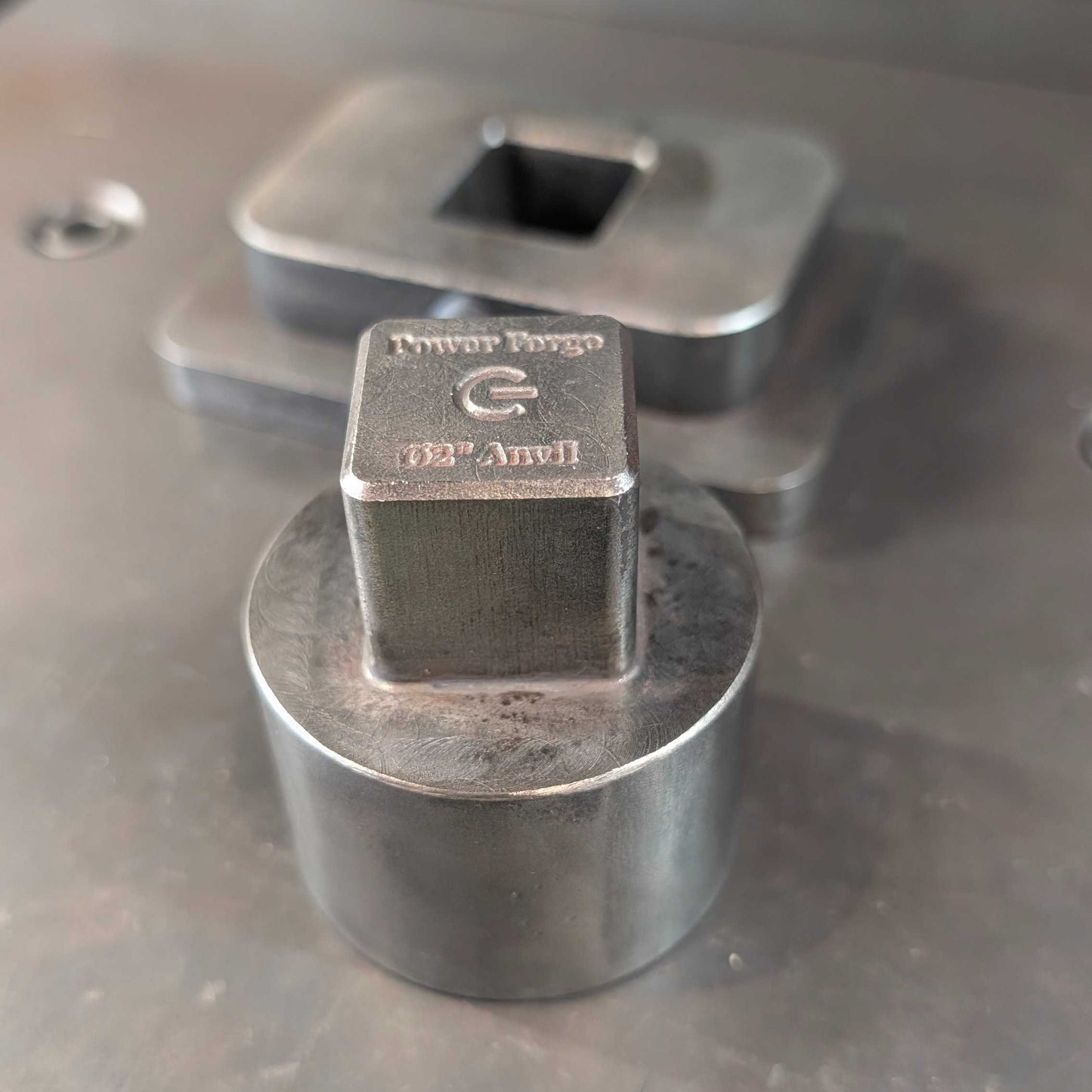 A close-up of a metallic cylindrical clamp or fixture with a square metal block on top, showing engraved text and a square recess on the top surface, set on a gray metal work surface.