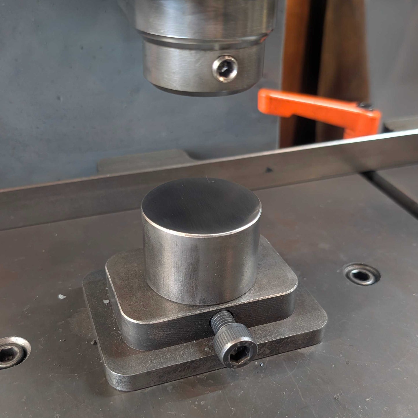 A close-up of a metallic cylindrical workpiece mounted on a square base on a metal machine table, with part of a press or milling machine visible above and an orange handle in the background.
