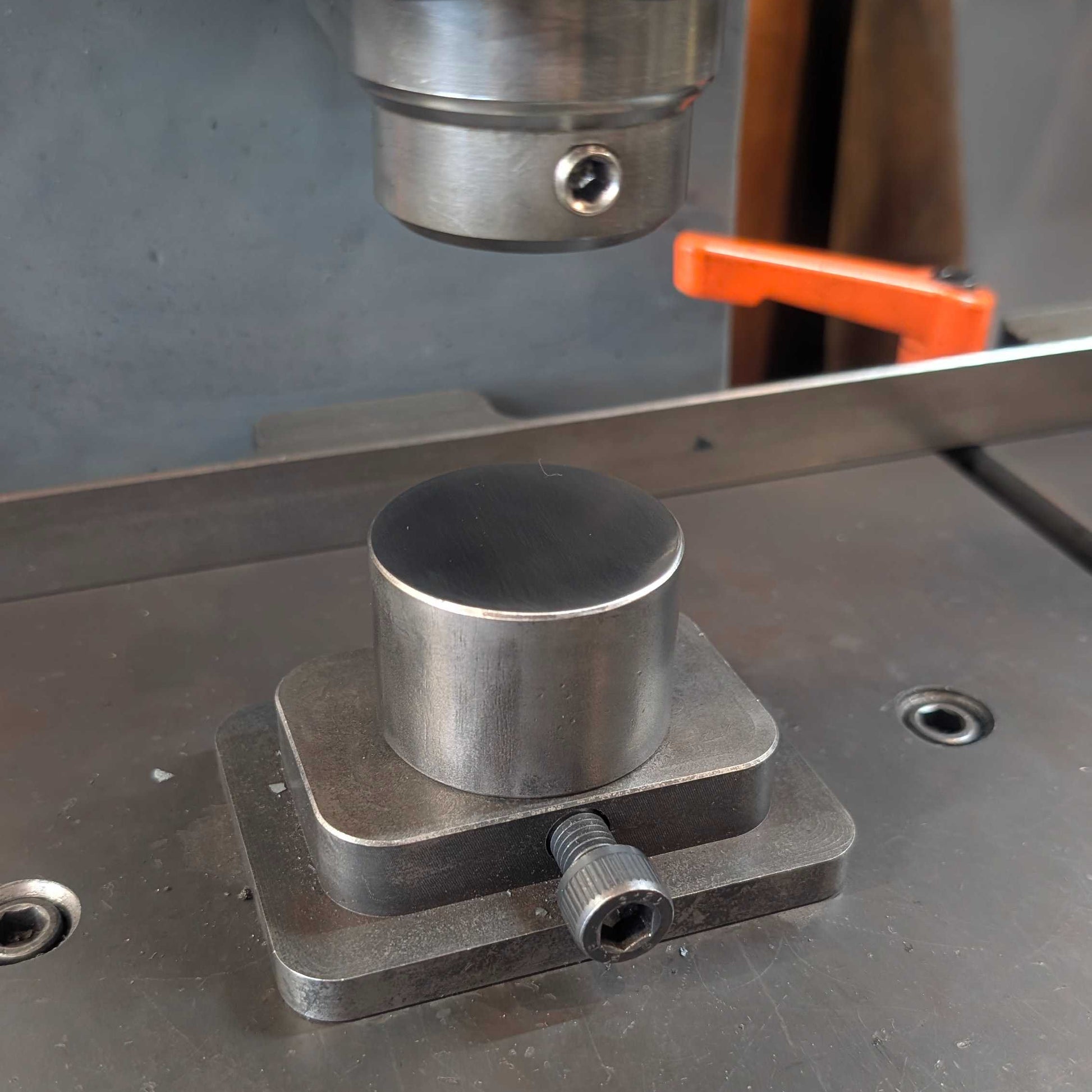 A close-up of a metallic cylindrical workpiece mounted on a square base on a metal machine table, with part of a press or milling machine visible above and an orange handle in the background.