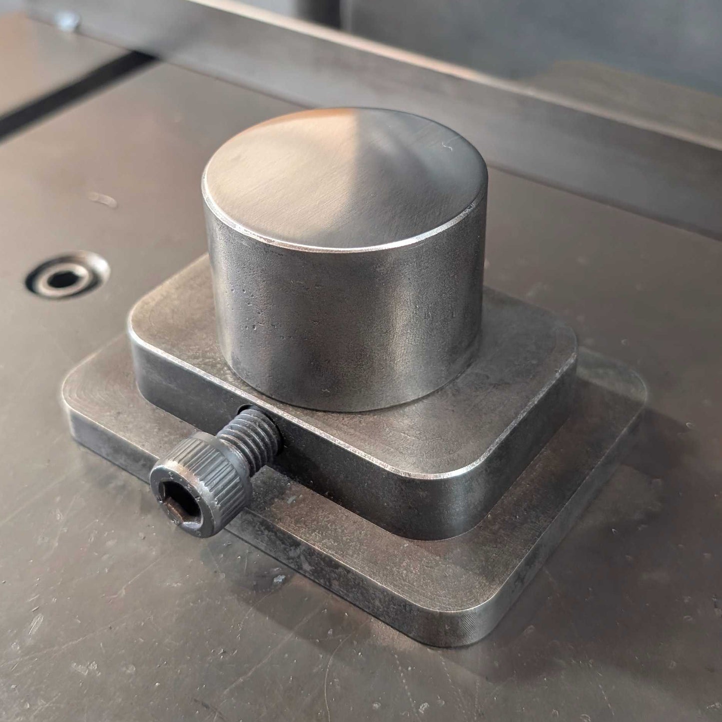 A close-up of a metallic cylindrical metal component mounted on a square base under a press or machine ram, with an orange-handled tool visible in the background on a metal work surface.