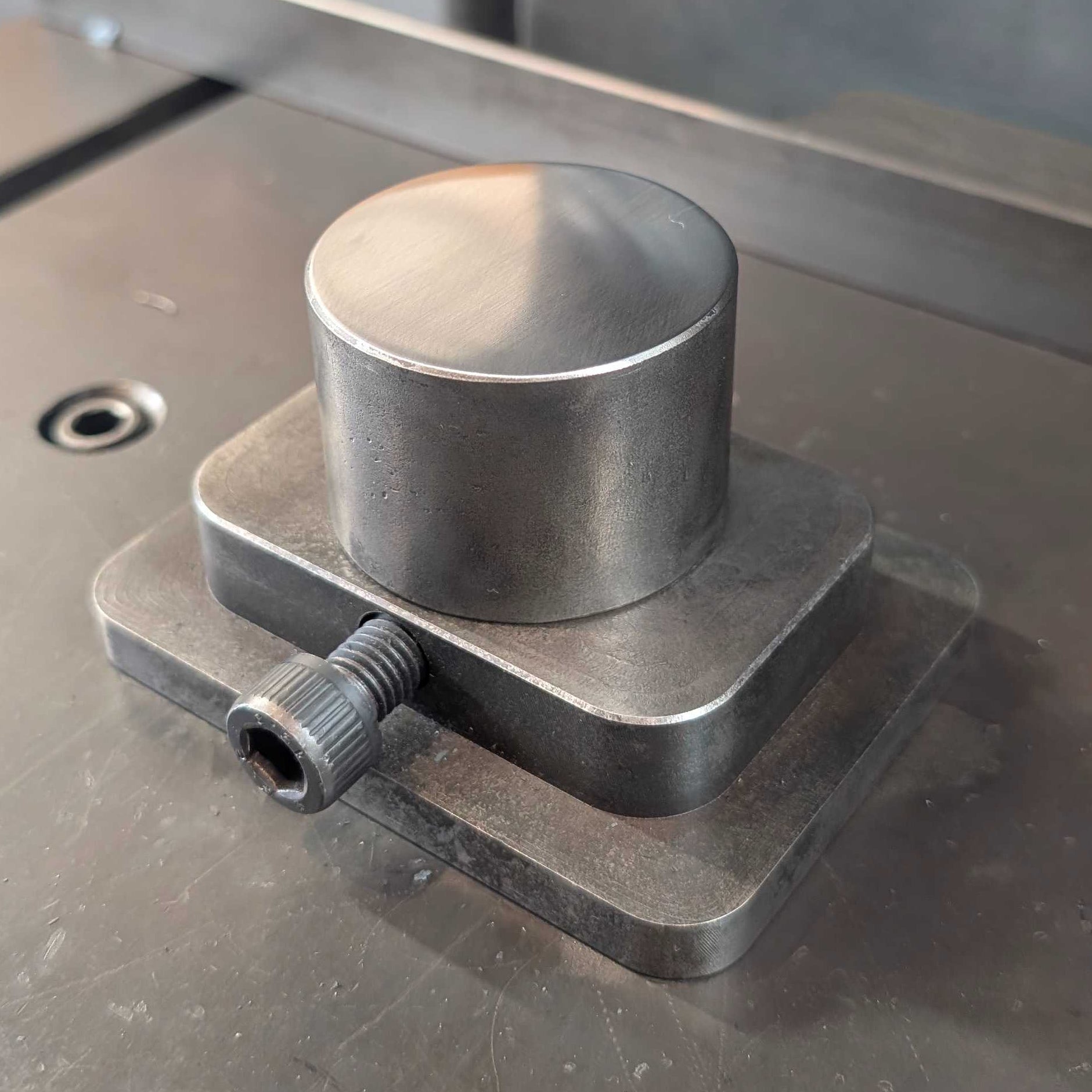 A close-up of a metallic cylindrical metal component mounted on a square base under a press or machine ram, with an orange-handled tool visible in the background on a metal work surface.