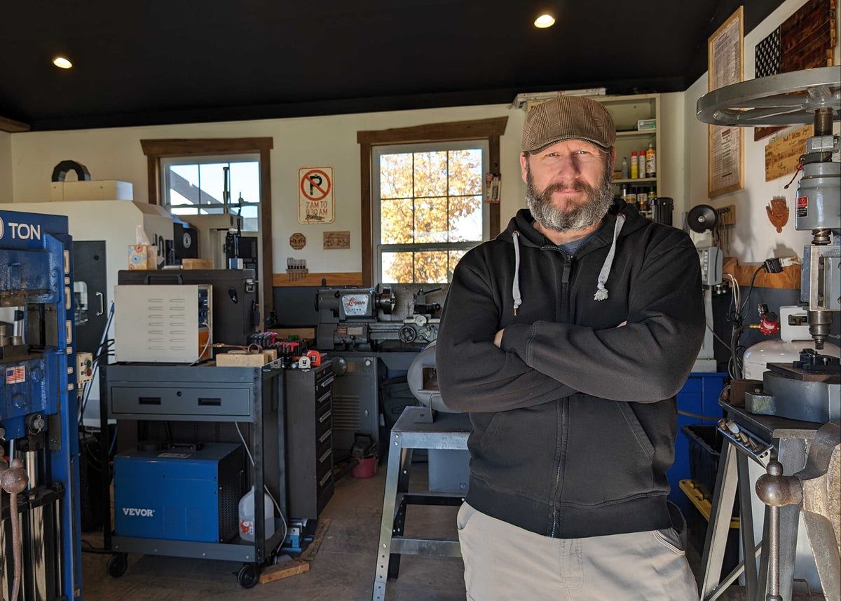 A man with a beard wearing a brown cap, black hoodie, and light beige pants stands with arms crossed inside a workshop filled with machinery and tools.