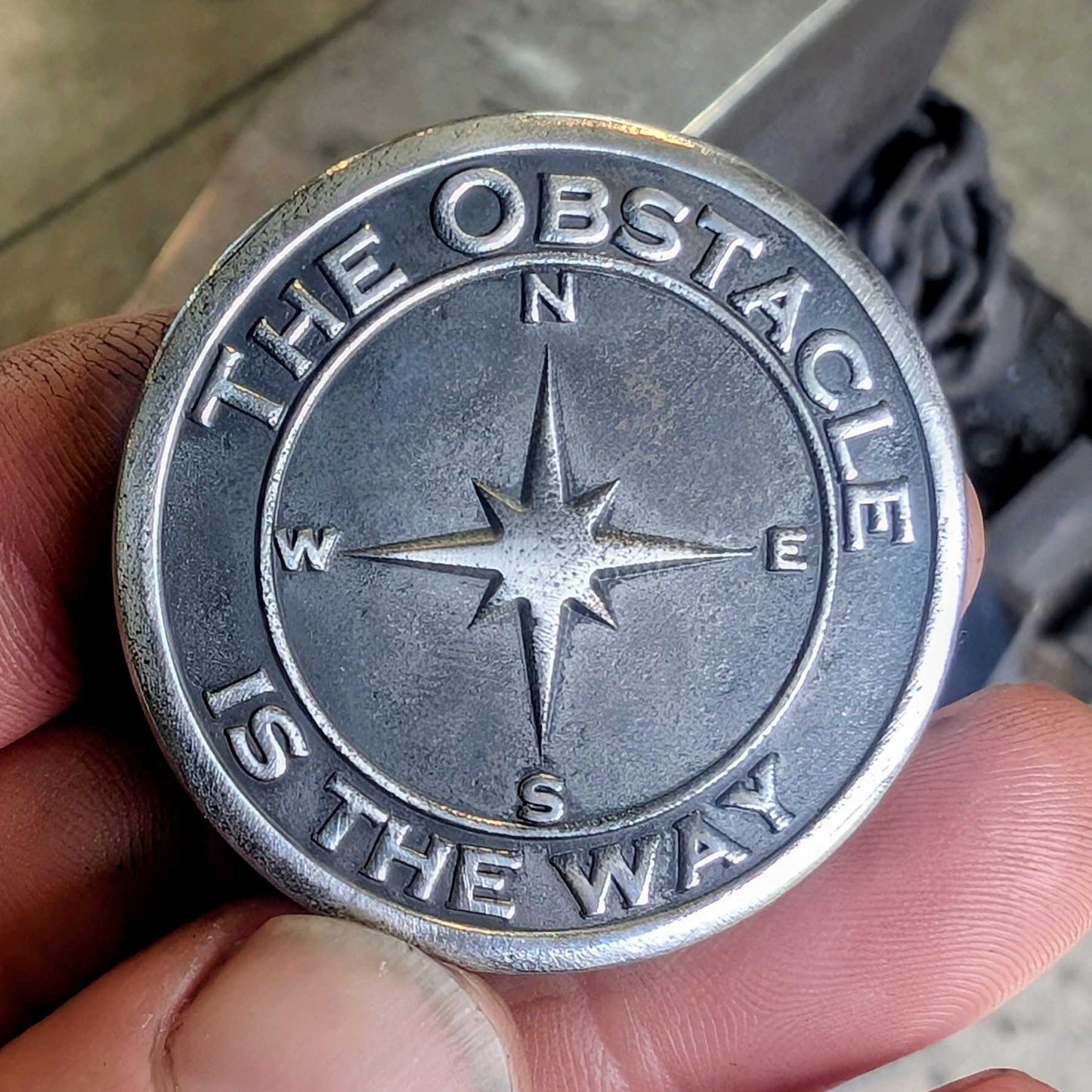 Metallic coin with 'The Obstacle is the Way' inscription held by a hand.