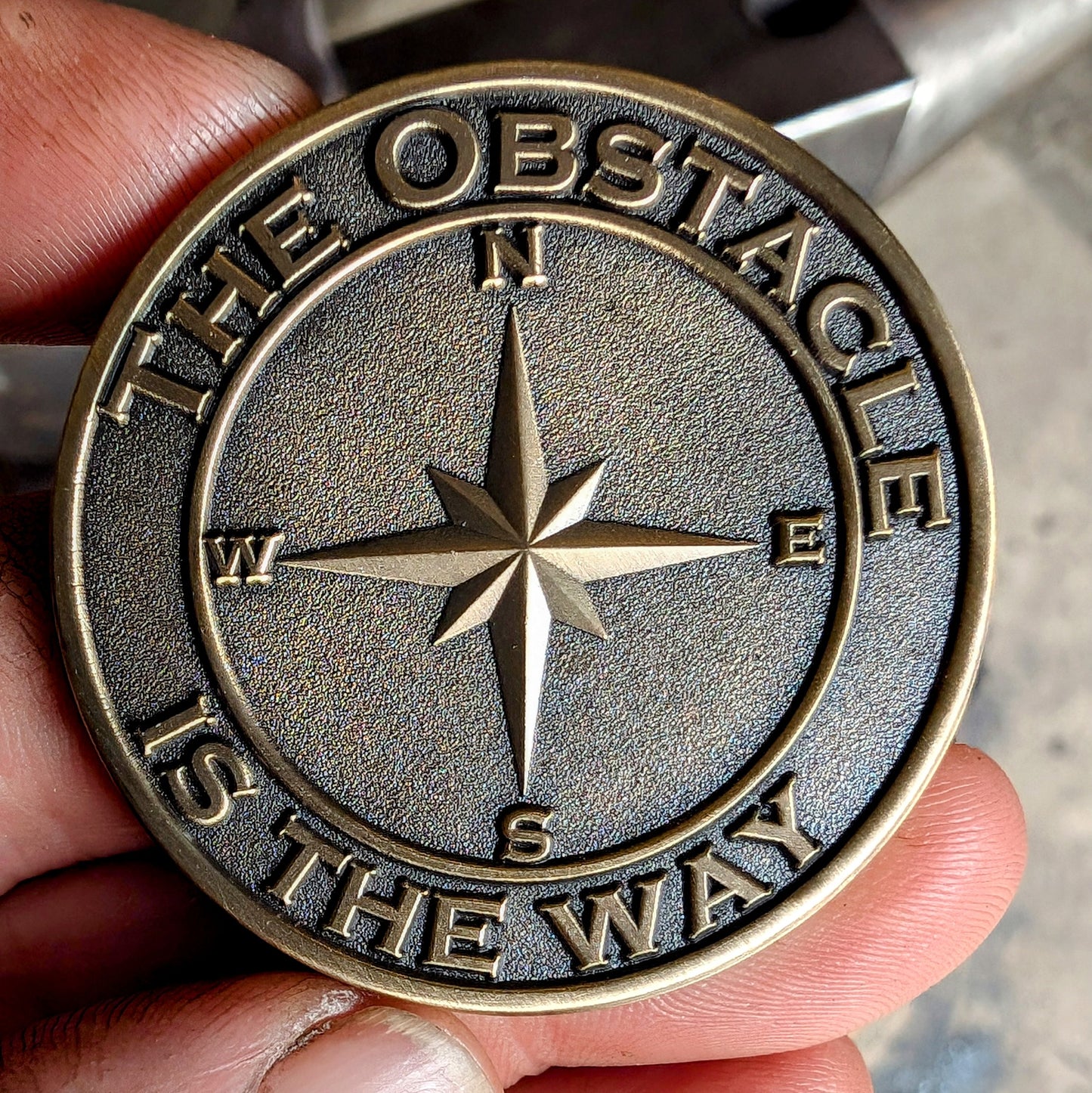 A bronze-finished solid brass coin with the inscriptions 'The Obstacle is the Way' and 'Embrace the Suck', along with a compass rose design.