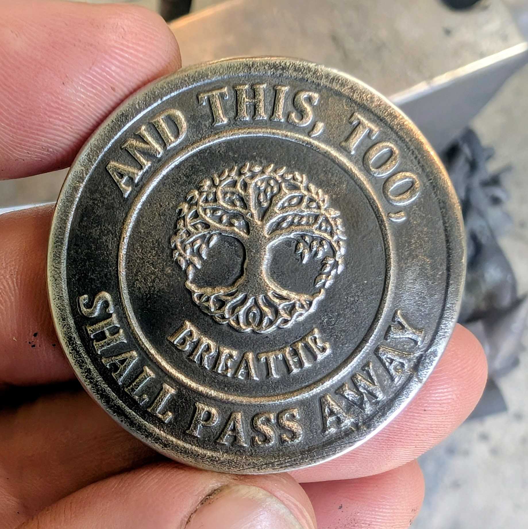A Stainless Steel Coin with the phrase And this, Too, Shall Pass Away surrounding the tree of life and the word breathe.