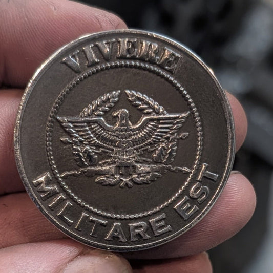 A close-up of a hand holding a round metallic coin or medallion with an eagle emblem and the text 'VIVERE MILITARE EST'.